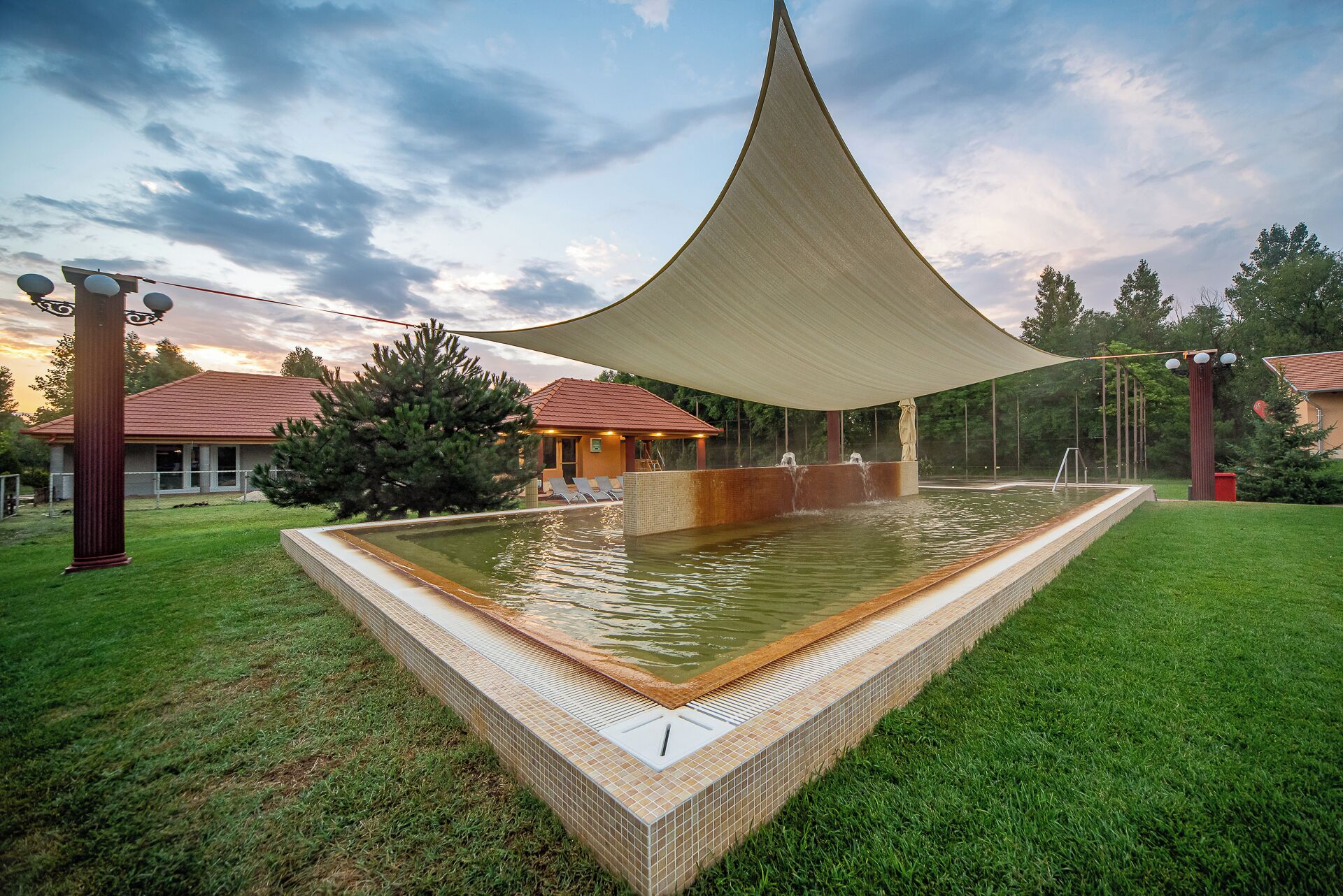 A swimming pool with a shaded area, surrounded by grass and trees, and a building with a red roof in the background.