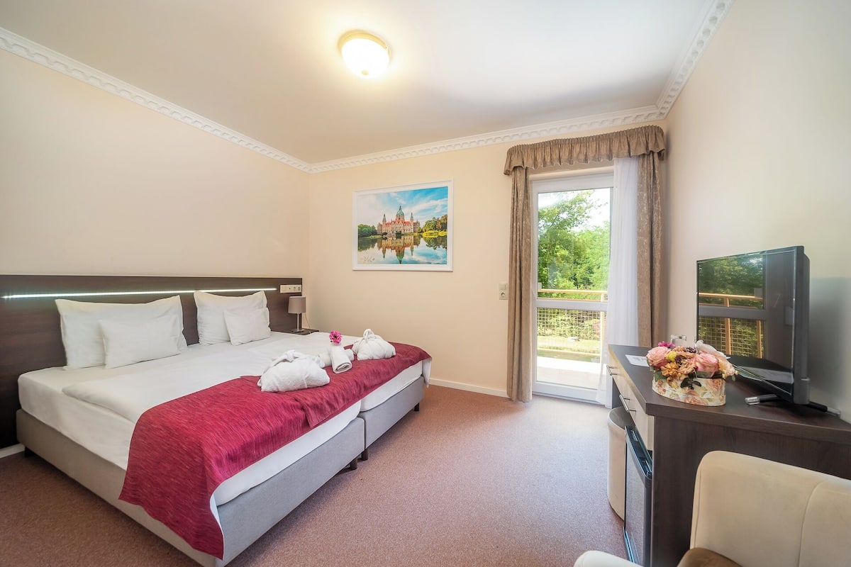 Habitación de hotel con cama, escritorio con televisor y ventana con vista a follaje.