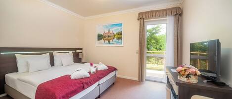 A hotel room with a bed, a desk with a television, and a window with a view of greenery.