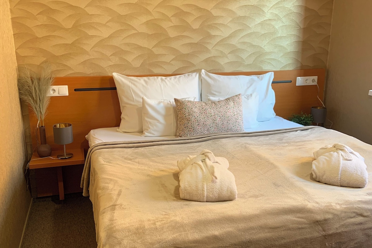 A hotel room with a bed, two pillows, a bedside table with a lamp, a wall air conditioner, and a red ottoman.