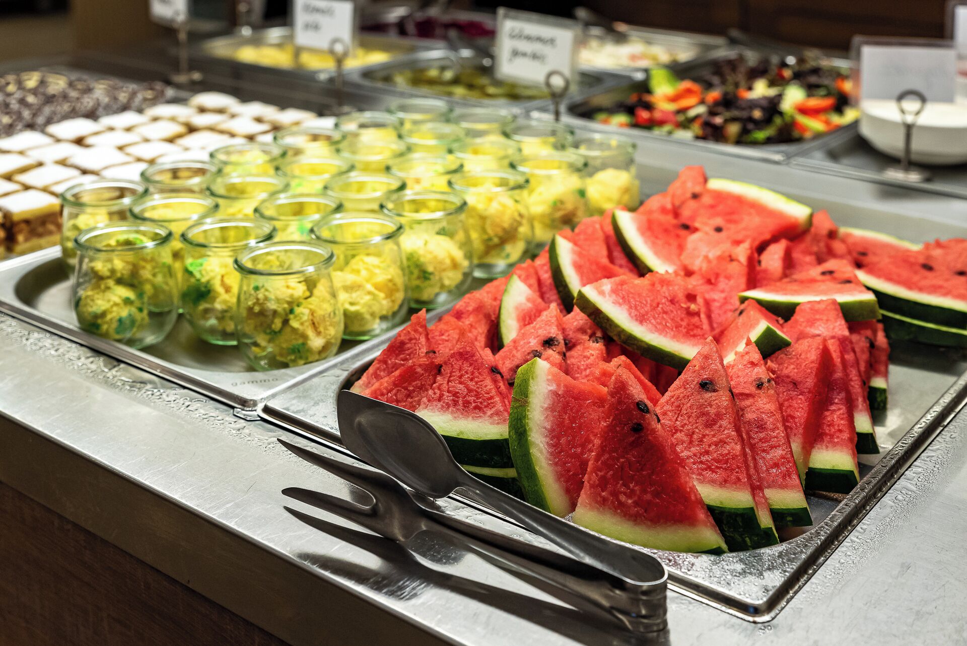 Un bufé con diversas opciones de comida, incluyendo sandía en rodajas y una selección de platos en recipientes de vidrio.