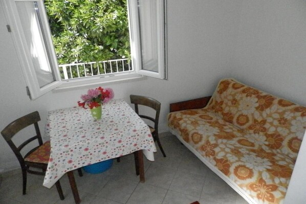Une petite pièce avec une table et des chaises, un canapé, une fenêtre donnant sur un paysage verdoyant et un vase rempli de fleurs posé sur la table.