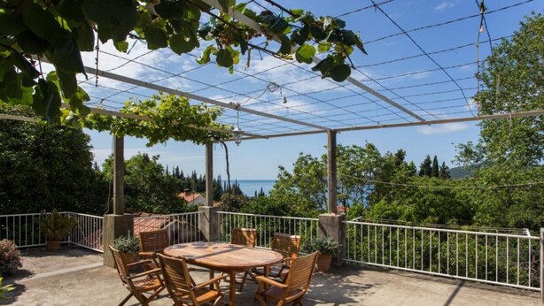 Un patio exterior con una mesa redonda y sillas, rodeado por una barandilla metálica blanca, con vistas panorámicas al mar y la vegetación.
