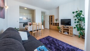 A modern living room with a grey sofa, a dining table with chairs, a TV on a wooden stand, and a kitchen in the background.