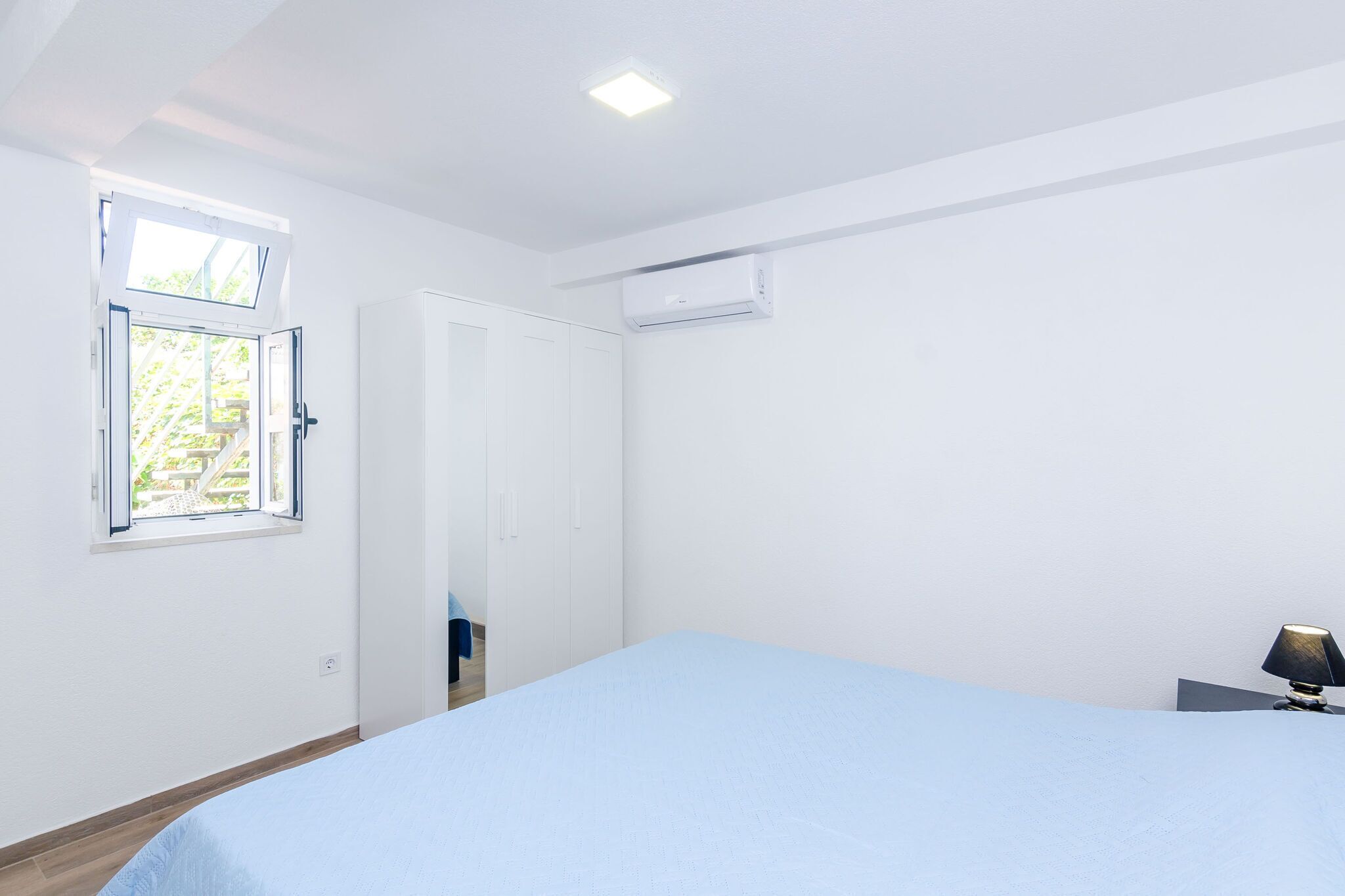 A bedroom with a bed, a window, an air conditioning unit, and a nightstand with a lamp.