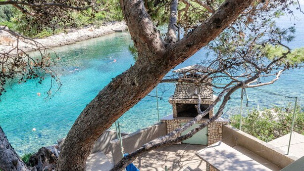 A stone-built outdoor seating area with a view of a clear blue body of water and a tree in the foreground. - Seafront Apartments Olivera-One Bedroom Apartment with Balcony and Sea View ( Ante ) (Blato)