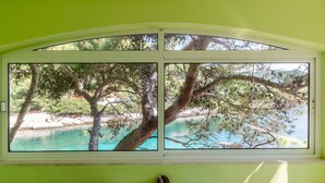 A room with a large window overlooking a turquoise body of water, surrounded by trees and a rocky shoreline. - Seafront Apartments Olivera-One Bedroom Apartment with Balcony and Sea View ( Ante ) (Blato)