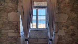 Una habitación con paredes de piedra y una ventana con cortinas color beige, con vista a una playa.