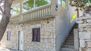 A stone building with a white door and brown shutters, a balcony with white balustrades, and a staircase leading up to a stone wall.