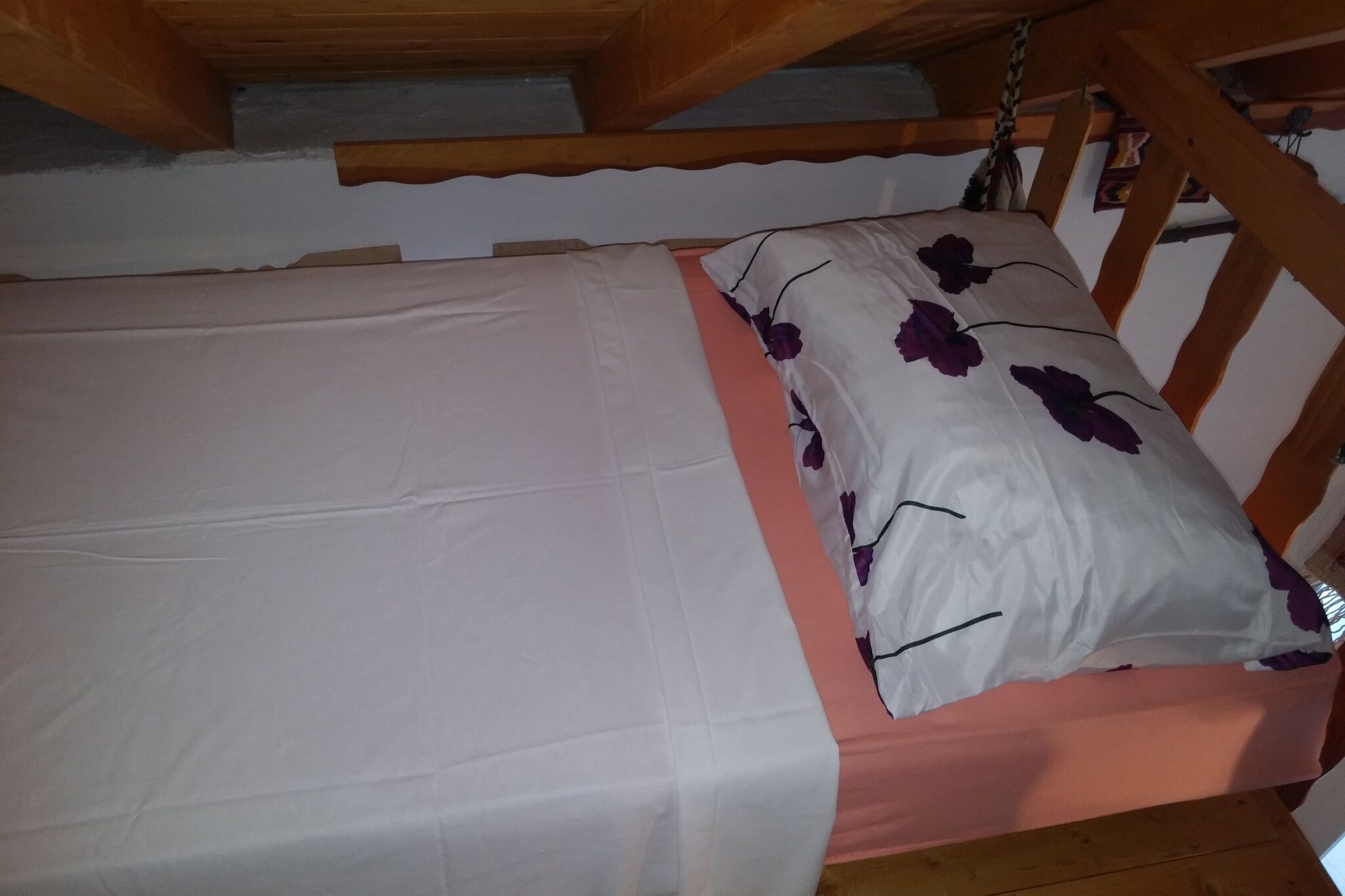 A single bed with a white sheet and a floral pillow, wooden headboard, and a wooden ceiling structure.