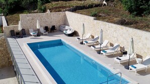 Una piscina rectangular rodeada por un muro de piedra, con sillas de descanso blancas y sombrillas.