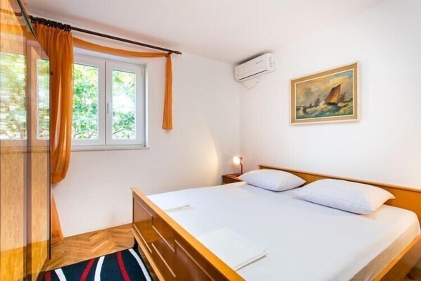 A hotel room with a bed, a nightstand, an air conditioning unit, a window with curtains, and a painting on the wall. - Guest House Raguz - Double Room - 2 (Dubrovnik)