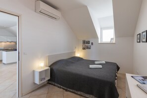 A bedroom with a bed, bedside lamps, a small bedside table, a wall-mounted air conditioner, and a window.
