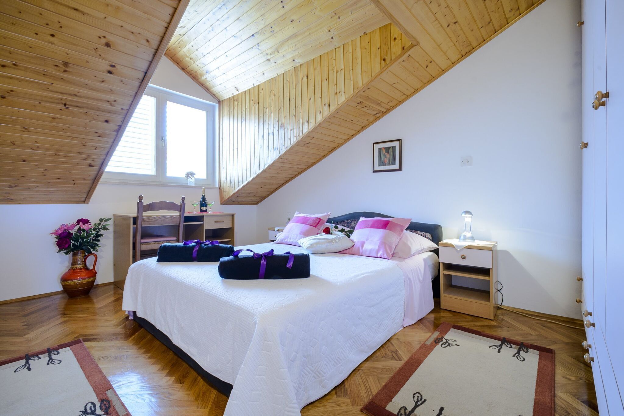 A bedroom with a sloped ceiling, a bed with white bedding and pink pillows, a bedside table with a lamp, and a wooden floor.