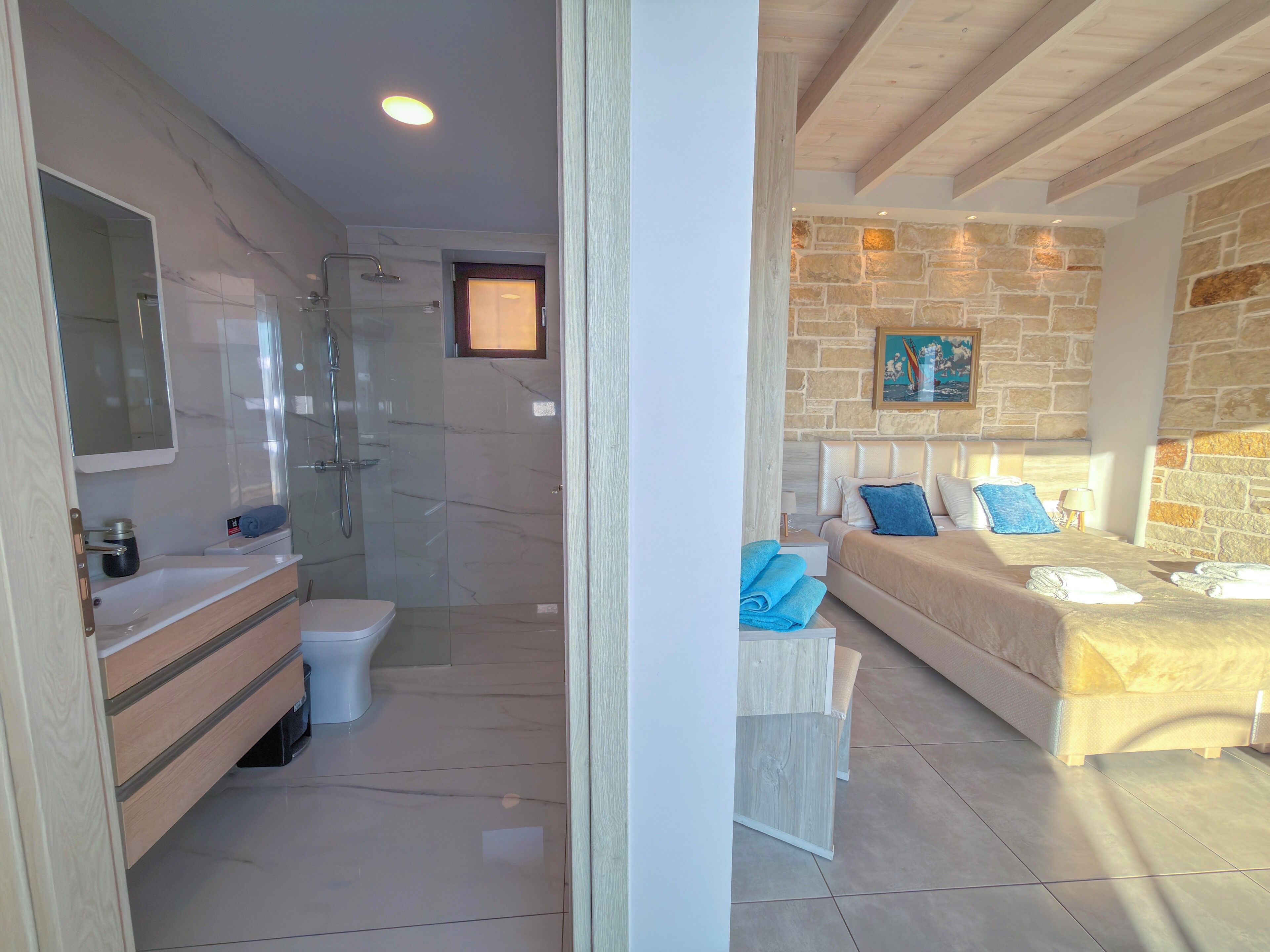 A modern bathroom with a shower, toilet, and vanity. There is a bed with blue towels and a stone wall in the adjacent room.