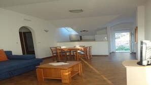 A spacious living area with a blue sofa, wooden coffee table, and dining table with chairs. A kitchen is visible in the background with a TV and a window.