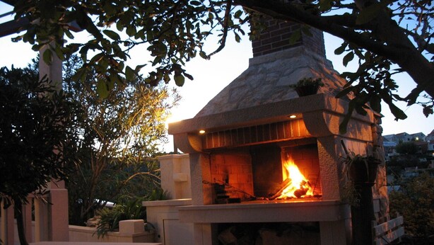 Chimenea de piedra al aire libre con fuego encendido, rodeada de una estructura pétrea y follaje.