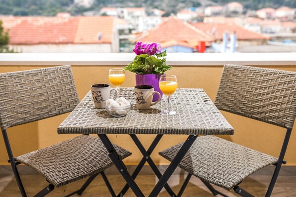 Een balkon met rieten meubels, een tafel met twee mokken, een vaas met bloemen en twee glazen jus d'orange.