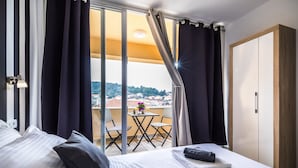 A hotel room with a bed, a small table with a vase and a remote control, a balcony with chairs, and a view of a cityscape. - Apartments & Rooms Sunshine Home - Double Room with balcony ( Black ) (Vela Luka)