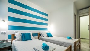 Une chambre d’hôtel avec deux lits, chacun agrémenté d’une serviette bleue et de draps blancs. On y trouve une table de chevet avec une lampe et une petite table. La chambre présente un design moderne, avec des rayures bleues et blanches sur le mur.