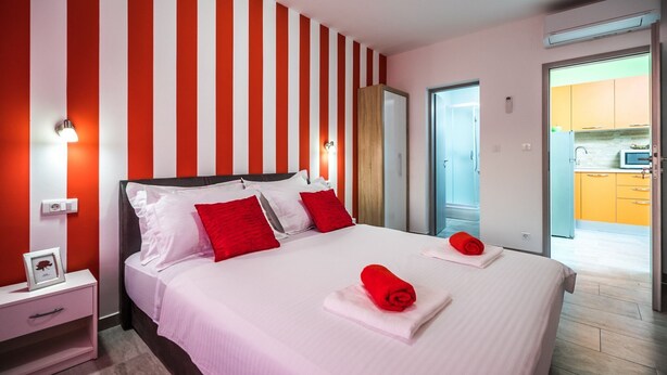 A hotel room with a large bed, red and white striped walls, a nightstand with a framed picture, and a view of the kitchen through an open door.