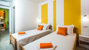 A hotel room with two single beds, each with a white bedspread and an orange pillow. The room has a green door leading to a kitchen area, and the walls are painted white with large yellow panels.