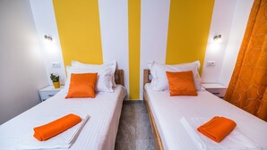 A hotel room with two beds, each with white linens and orange pillows. There are nightstands with lamps and a small plant on one nightstand.