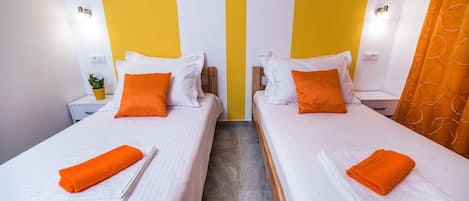 A hotel room with two beds, each with white linens and orange pillows. There are nightstands with lamps and a small plant on one nightstand.