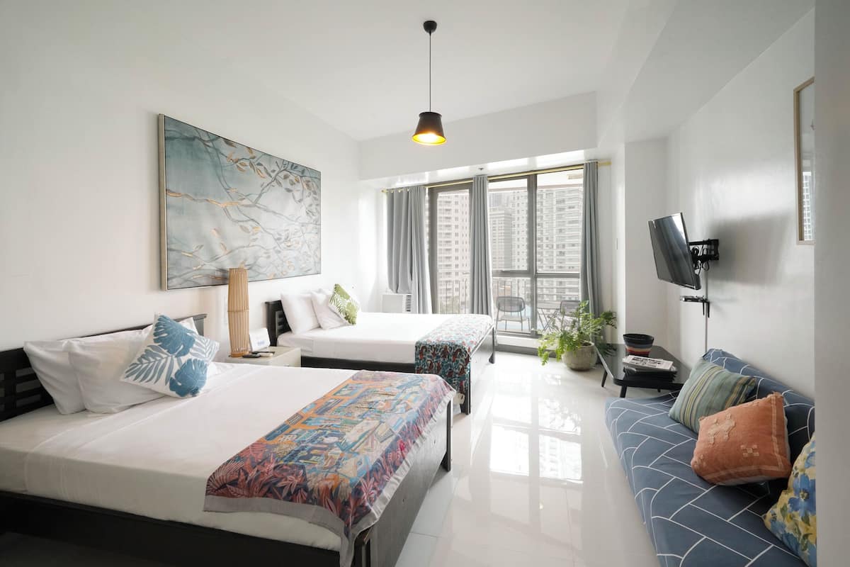 A modern hotel room with two beds, a sofa, a TV, and a large window with a city view.