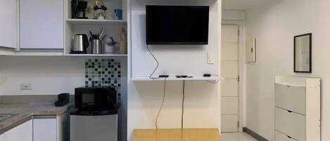 Ett modernt kök med vita skåp, en inbyggd mikrovågsugn, ett litet kylskåp, en träbänk och en platt-TV som är monterad på väggen.