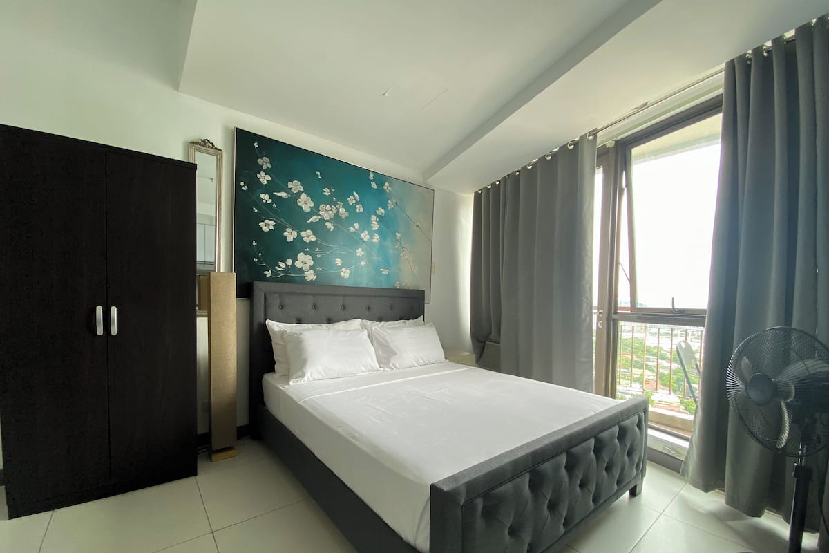 A bedroom with a bed, a wardrobe, a painting, a window with curtains, and a standing fan.