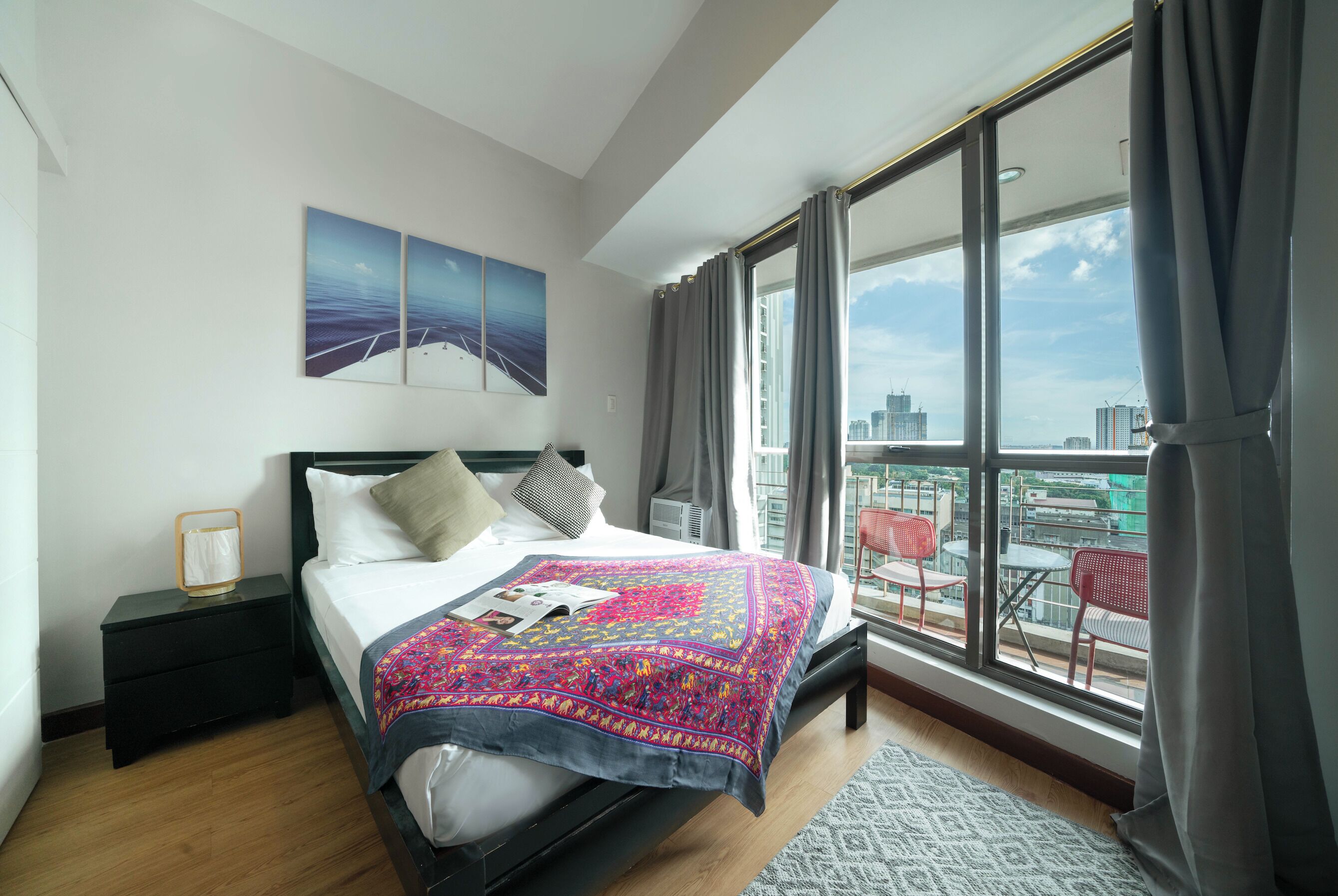 A modern hotel room with a large bed, a nightstand, a window with a city view, and a balcony with outdoor seating.