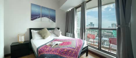 A modern hotel room with a large bed, a nightstand, a window with a city view, and a balcony with outdoor seating.