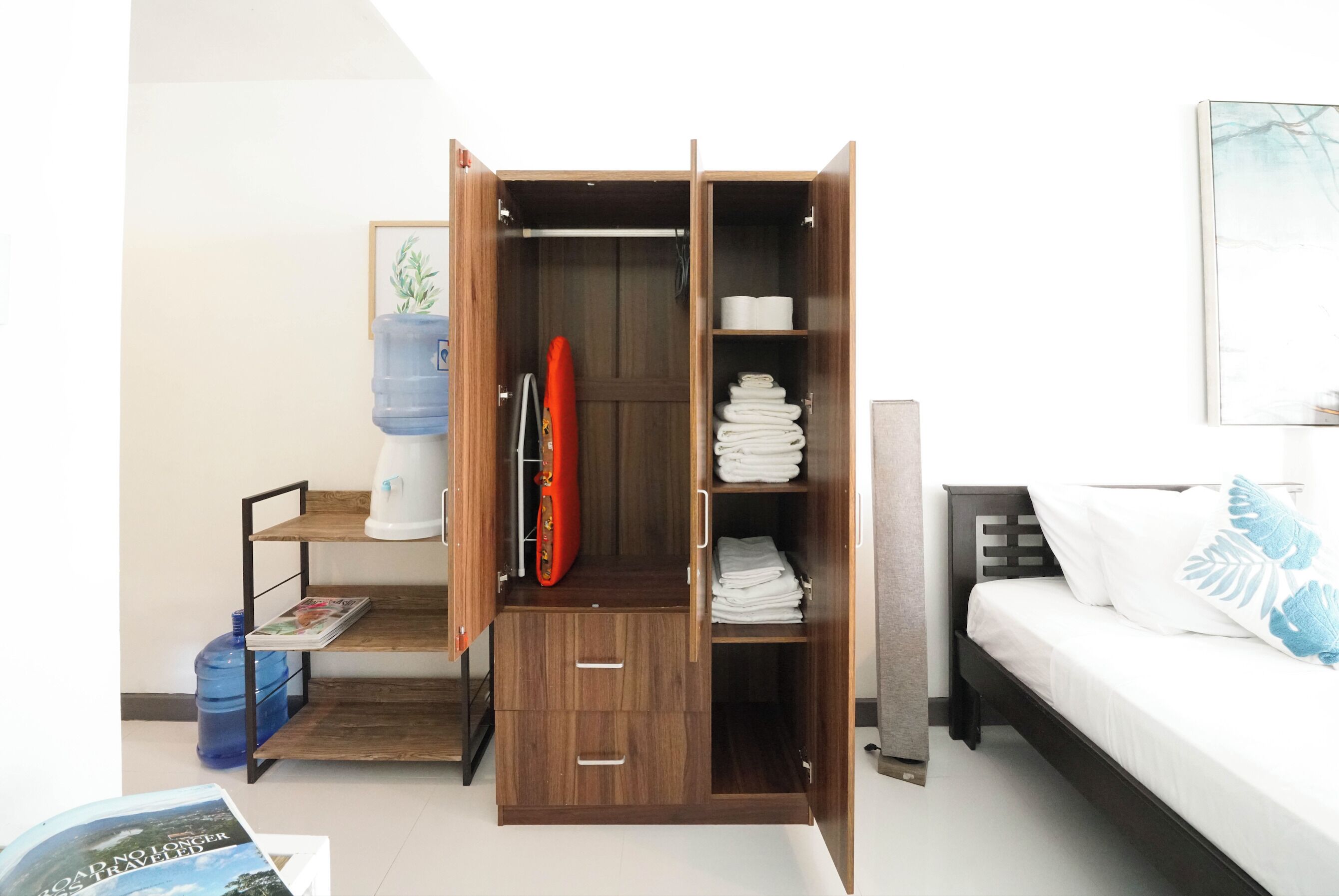Une chambre à coucher comprenant un lit, une armoire avec plusieurs compartiments, un distributeur d’eau et une petite étagère avec un magazine.