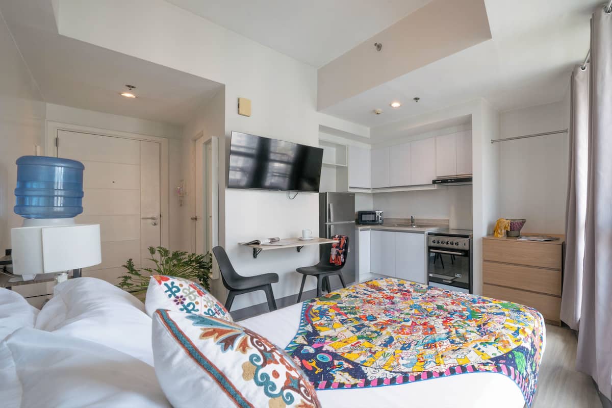 A compact hotel room with a bed, a desk, a kitchenette, and a large water dispenser.