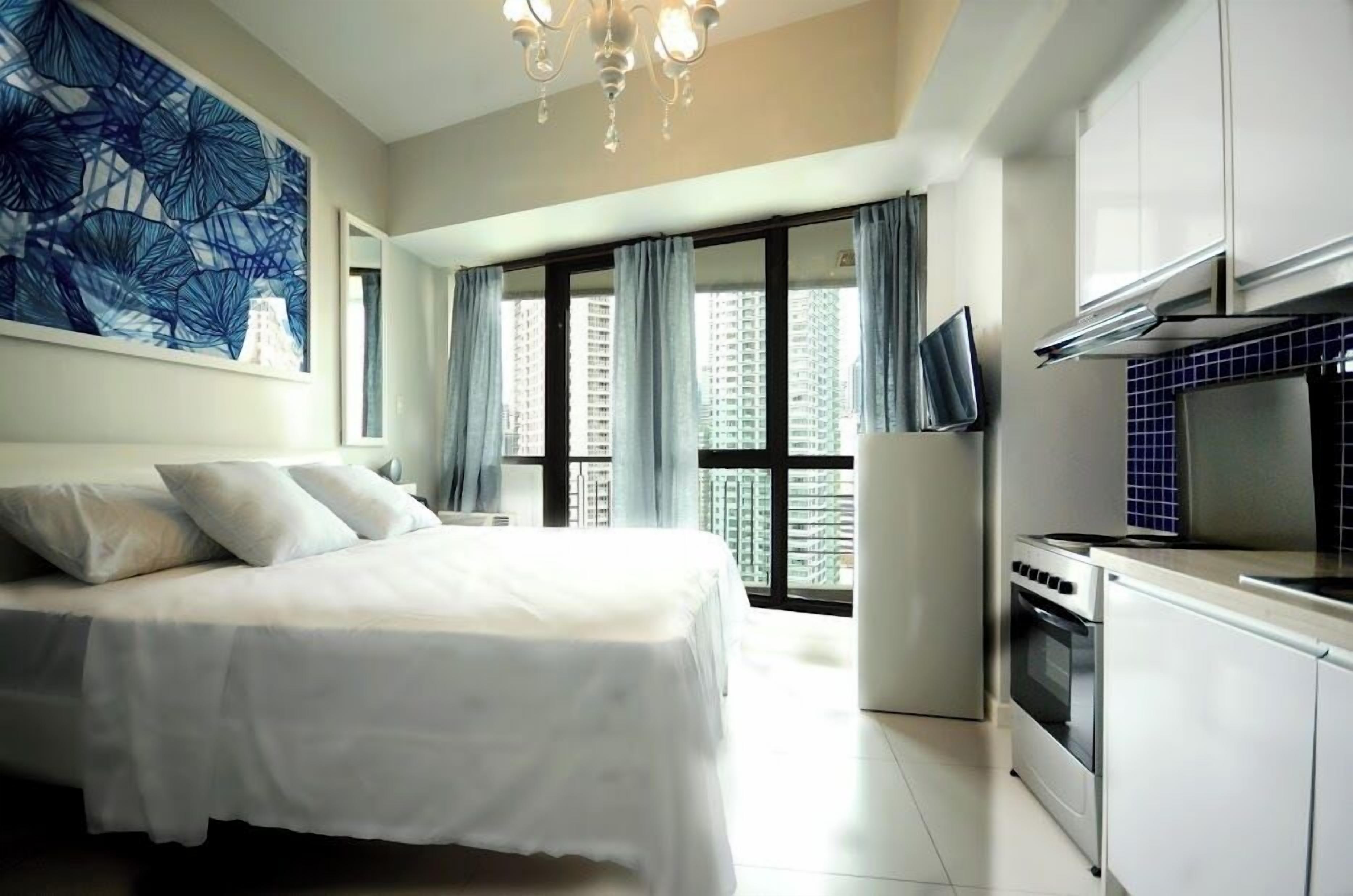A modern bedroom with a large bed, a TV, a kitchenette, and a view of high-rise buildings.