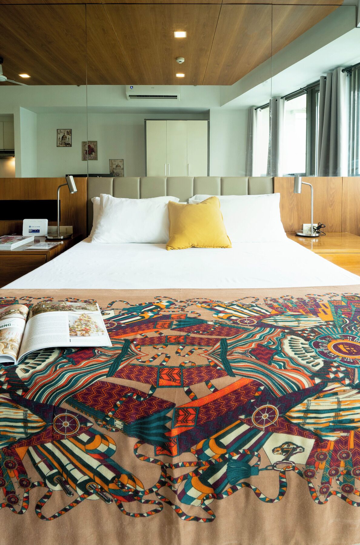 A hotel room with a large bed, a wooden ceiling, a headboard, a bedside table with a lamp, a window with curtains, and a wall-mounted air conditioner.