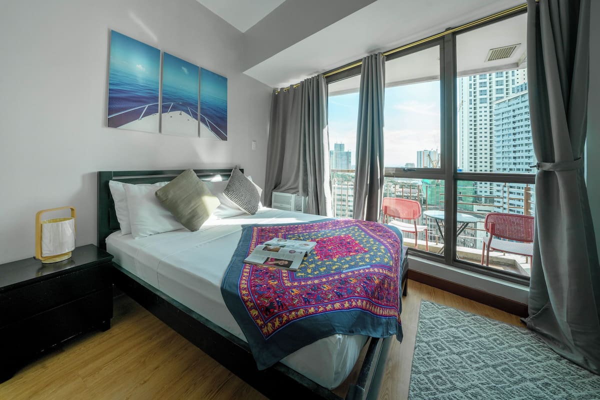 A modern hotel room with a large bed, a view of the city through floor-to-ceiling windows, and a balcony with outdoor seating.