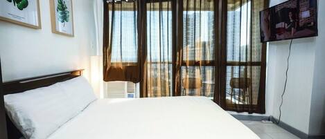 Habitación de hotel con una cama grande, una lámpara en el techo, un televisor montado en la pared y una ventana con cortinas.