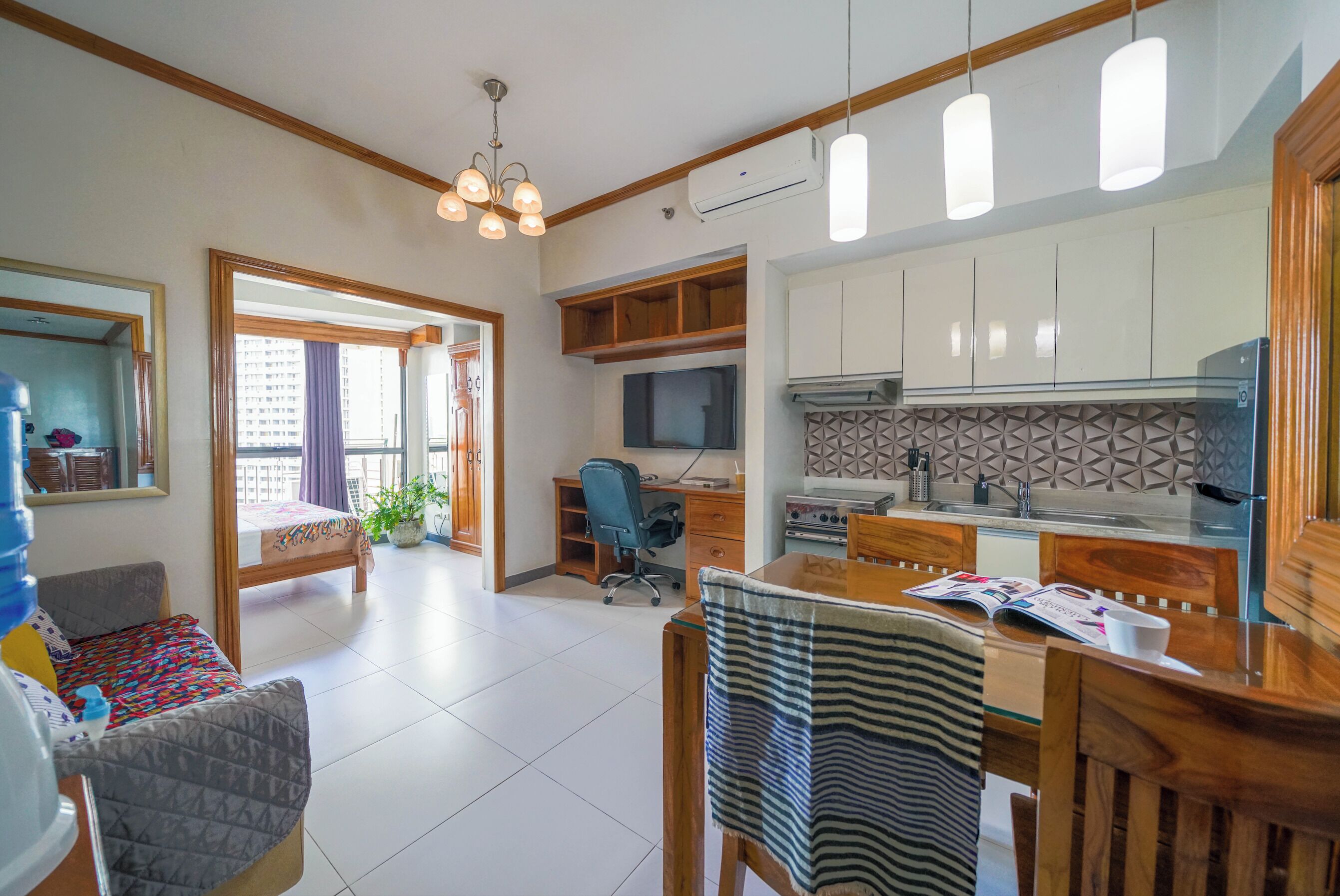 A modern kitchen with white cabinets, a built-in oven, and a microwave. There is a dining area with a wooden table and chairs, and a small seating area with a sofa and a water dispenser.
