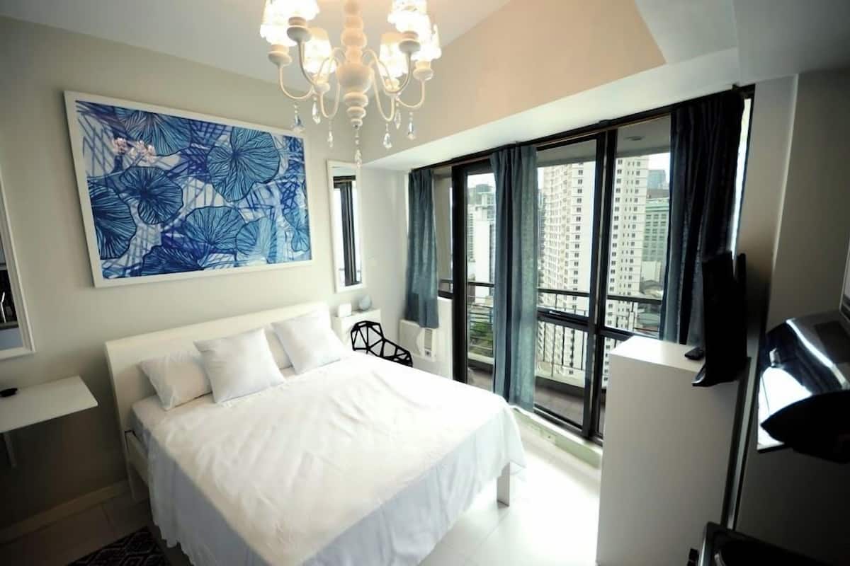A modern hotel room with a large bed, a chandelier, a city view through large windows, and a blue artwork on the wall.