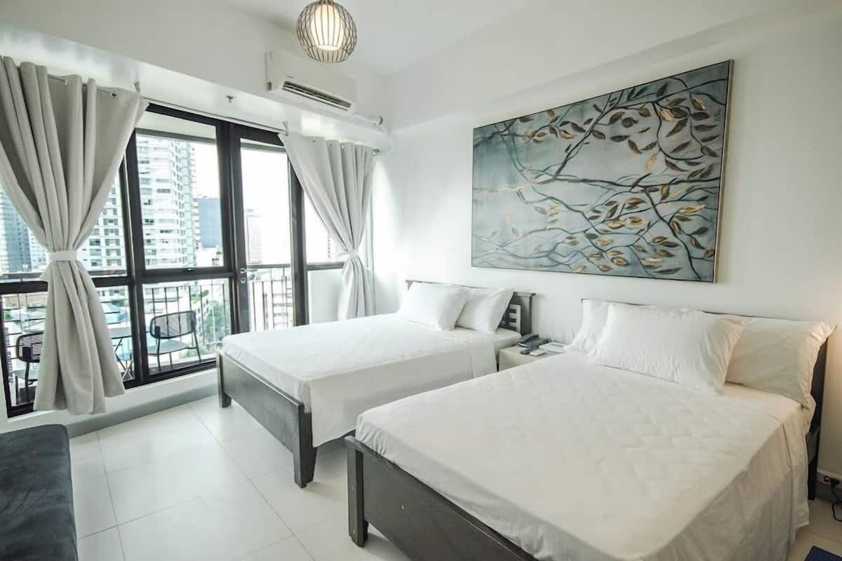 A hotel room with two beds, a large window with curtains, a ceiling light, and a wall-mounted painting.