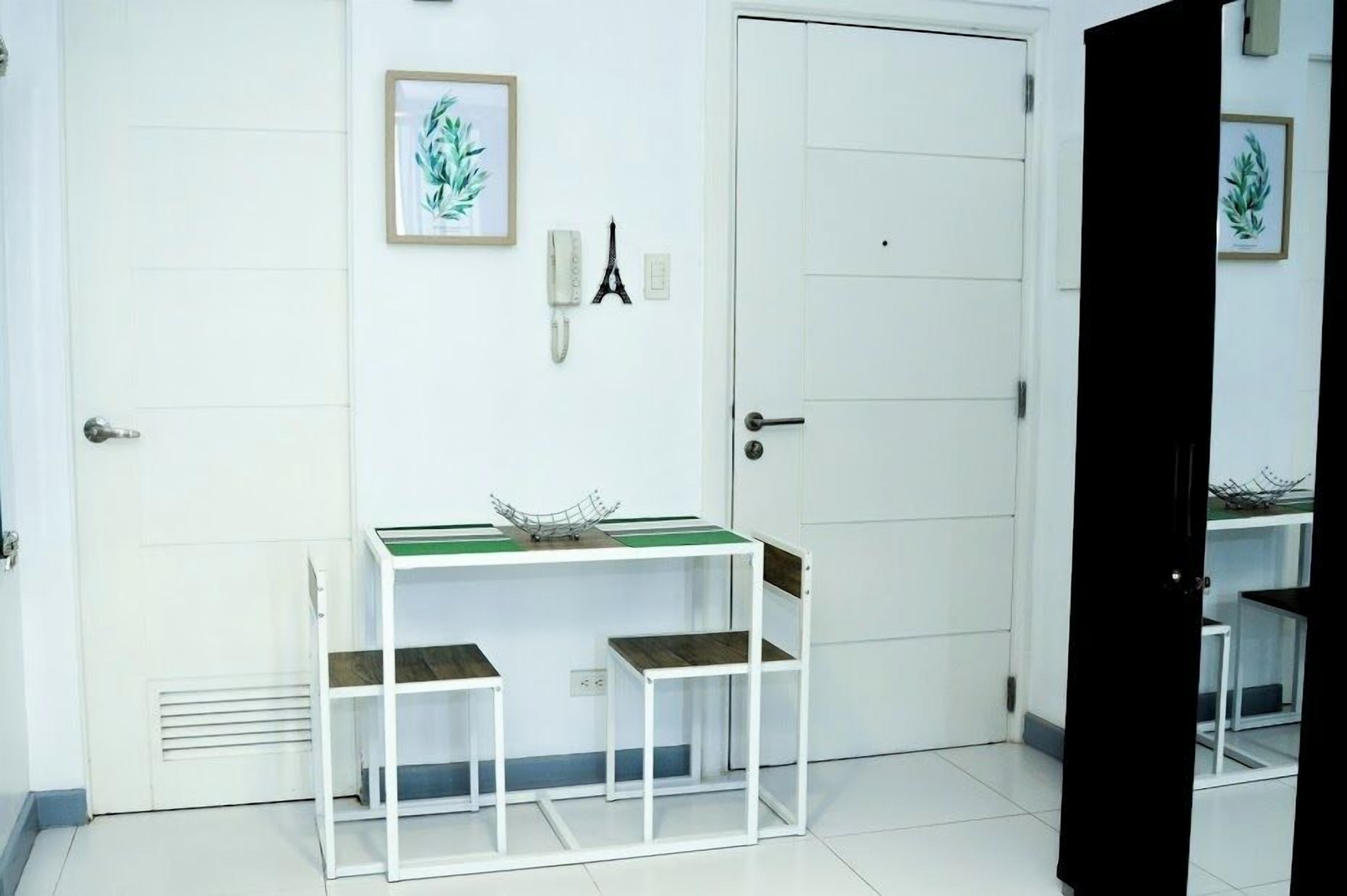 Une pièce avec une porte blanche, une petite table à plateau vert, deux chaises en bois, une étagère blanche et un tableau encadré accroché au mur.