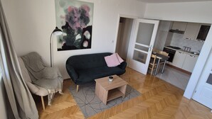 A modern living room with a black sofa, a small wooden coffee table, and a painting on the wall. A chair with a checkered blanket is visible, and a kitchen area is seen through an open door.