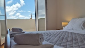 A hotel room with a bed, a window with a view of the sky and a distant structure, and a nightstand with a lamp.