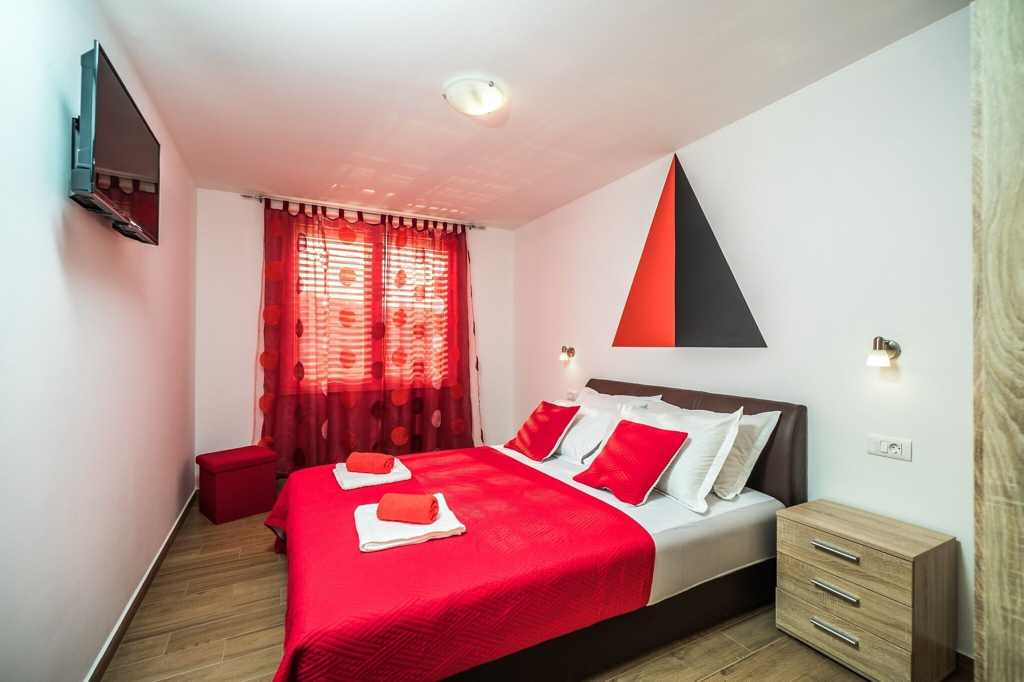 Una camera da letto moderna con un letto grande, biancheria rossa, un comodino in legno e una TV a parete.