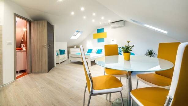 A modern dining area with a glass table and yellow chairs, a white sofa with blue cushions, and a skylight above. - Apartments & Rooms Sunshine Home - Studio Apartment (Vela Luka)