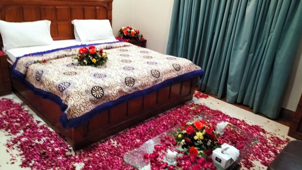A neatly made bed with a patterned bedspread, a small table with a vase of flowers, and a carpet of rose petals. - Cosy inn Near Millennium Mall  (Karachi)