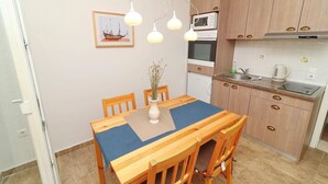 Een compacte keuken met houten kastjes, een inbouwoven en een magnetron. Er staat een eettafel met stoelen en een vaas met gedroogde planten.