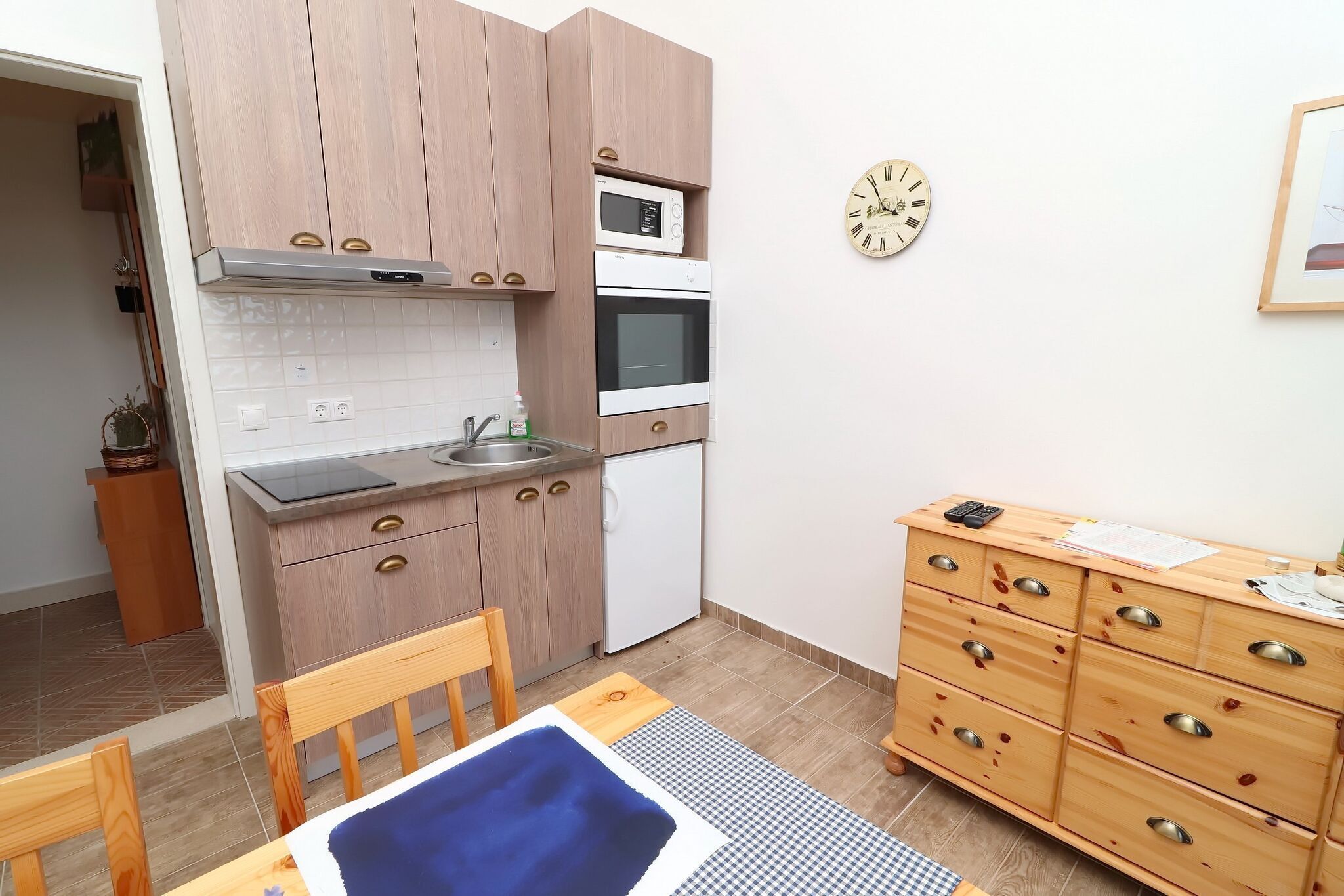 Une cuisine compacte avec des meubles en bois, un four à micro-ondes, un four traditionnel et un réfrigérateur. Il y a une horloge au mur et une petite table avec une chaise.